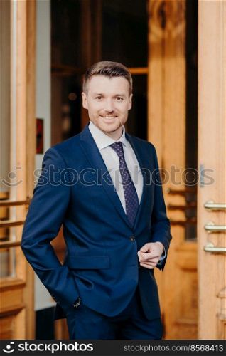 Attractive male executive manager in formal outft, keeps hand in pocket, has positive expression, stands near office, rejoices good achievement, confident in success. Male lawyer stands near doors