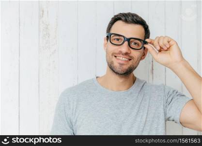 Attractive male model with stubble, looks joyfully through spectacles, has pleasant conversation with someone, wears casual t shirt, poses against white wooden background. Lifestyle concept.