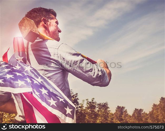 Attractive man holding a sledgehammer and a US Flag in his hands and looking into the distance against a background of trees, blue sky and the rays of the setting sun. National holiday concept. Attractive man holding a hammer and US Flag