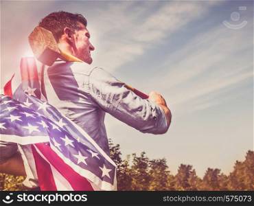 Attractive man holding a sledgehammer and a US Flag in his hands and looking into the distance against a background of trees, blue sky and the rays of the setting sun. National holiday concept. Attractive man holding a hammer and US Flag