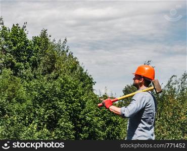 Attractive man holding a sledgehammer in the park against the backdrop of green trees. Close-up. Concept of labor and employment. Man holding a hammer in the park