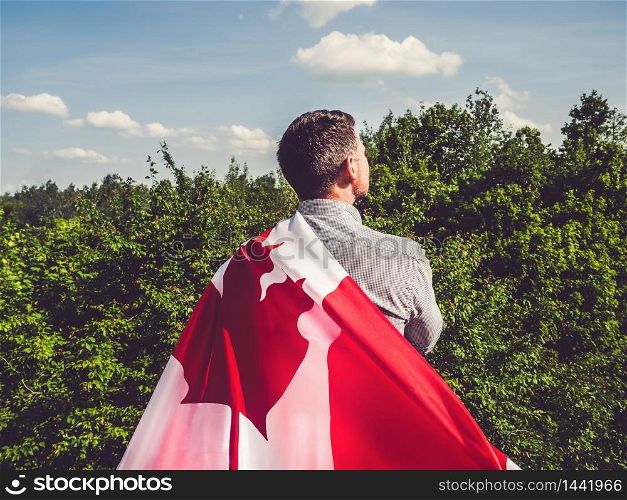 Attractive man holding Canadian Flag on blue sky background on a clear, sunny day. View from the back, close-up. National holiday concept. Attractive man holding Canadian Flag. National holiday
