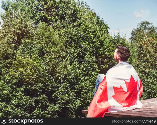 Attractive man in jeans and denim shirt holding a Canadian Flag against a clear, sunny, blue sky. View from the back, close-up. National holiday concept. Man holding a Canadian Flag. National holiday