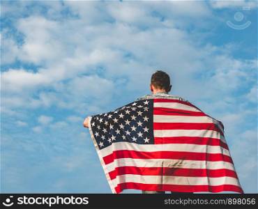 Attractive man in jeans and denim shirt waving an American Flag against a clear, sunny, blue sky. View from the back, close-up. National holiday concept. Handsome, young man waving an American flag