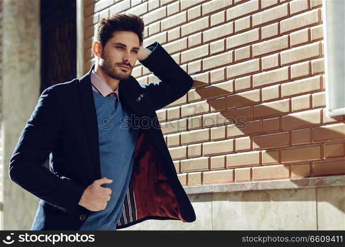 Attractive man in the street wearing british elegant suit. Young bearded businessman with modern hairstyle in urban background.