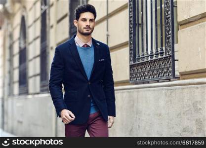 Attractive man in the street wearing british elegant suit. Young bearded businessman with modern hairstyle in urban background.