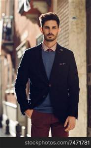 Attractive man in the street wearing british elegant suit. Young bearded businessman with modern hairstyle in urban background.. Attractive man wearing british elegant suit in the street. Modern hairstyle.