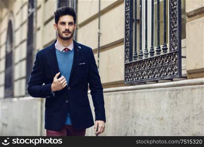 Attractive man in the street wearing british elegant suit. Young bearded businessman with modern hairstyle in urban background.. Attractive man wearing british elegant suit in the street. Modern hairstyle.