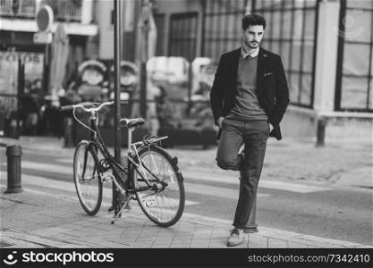 Attractive man in the street wearing british elegant suit near a vintage bicycle. Young bearded businessman with modern hairstyle in urban background.. Man wearing british elegant suit in the street near an old bycicle