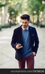 Attractive man in the street wearing british elegant suit with smart phone in his hand. Young bearded businessman with modern hairstyle in urban background.. Man in the street in formalwear with smartphone in his hand.