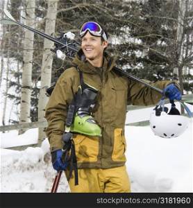 Attractive man in winter clothing walking in snow carrying ski equipment and smiling.