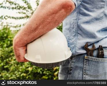 Attractive man in work clothes, holding construction helmet in his hands against the background of trees, blue sky and sunset. View from the back. Labor and employment concept. Attractive man holding construction helmet in his hands