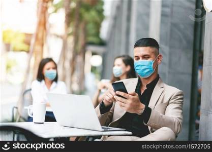 Attractive man texting message on mobile phone while sitting at outside of modern cafe. Businessman in face mask using smartphone while pandemic covid-19 virus. Business People in New normal Lifestyle