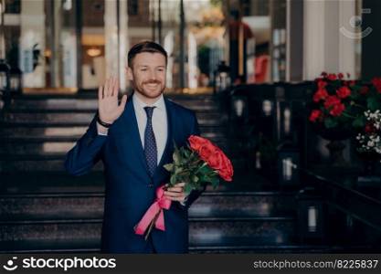 Attractive man waiting on street for date in elegant dark blue suit holding red rose flower bouquet next to restaurant outside while raising one hand in greeting romantic manner. Attractive man waiting on street in elegant dark blue suit with red rose flower bouquet