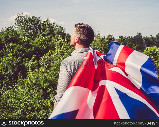 Attractive man waving a British Flag against a background of trees and blue sky. View from the back, close-up. National holiday concept. Attractive, young man waving a British Flag