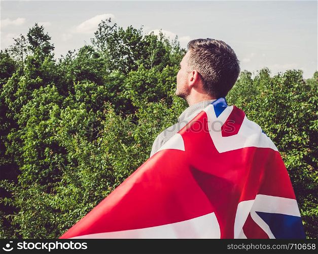 Attractive man waving a British Flag against a background of trees and blue sky. View from the back, close-up. National holiday concept. Attractive, young man waving a British Flag