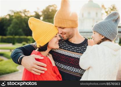 Attractive man wears yellow warm hat, embraces his wife and daughter, looks at them with great love. Adorable little girl feels support from parents, smiles happily while being on father`s hands