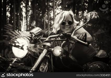 Attractive mature blonde woman biker in round dark eyeglasses clothing hippie style sitting on a motorcycle with the lights on in the forest. Monochrome, sepia filter.. Mature Woman Biker On a Motorcycle In Sepia