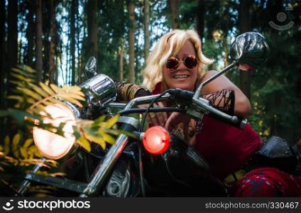 Attractive mature blonde woman biker in round dark eyeglasses clothing hippie style sitting on a motorcycle with the lights on in the forest.. Mature Woman Biker On a Motorcycle