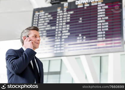 Attractive mature businessman talking on smartphone while standing against flight screen display in airport