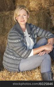 Attractive middle aged blond woman sitting on a hay bale in a barn wearing boots