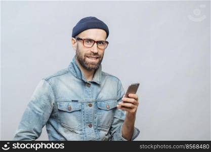 Attractive middle aged male model with bristle dressed in fashionable clothing, holds smart phone, chats with colleagues or friends in socail networks, isolated over white background. Technology