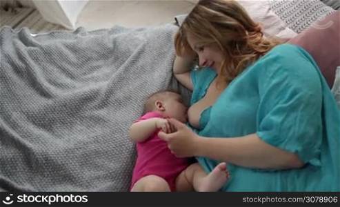 Attractive mother holding and caressing tiny fingers of her infant child while breastfeeding in bedroom. Caring mom nursing her baby girl, feeding breast to her newborn child. Top angle view. Slow motion. Steadicam stabilized shot.