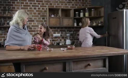 Attractive mother taking homemade cookies from the fridge and giving her happy daughter and grandmother in modern domestic kitchen. Overjoyed little girl and smiling grandmom awaiting to taste fresh handmade cookies at home. Dolly shot. Slow motion.