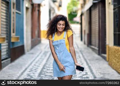 Attractive North African woman listening to music with earphones outdoors. Arab girl in casual clothes with curly hairstyle in urban background.