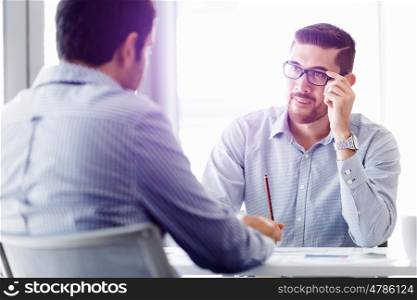 Attractive office worker sitting at desk. Young businessman sitting at desk in office and talking to his colleague