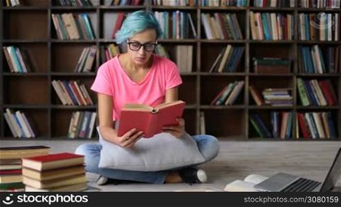 Attractive overwoked college female student with blue hair reading a book while studying hard in university library. Frustrated hipster girl putting eyeglasses on her head and massaging her nose bridge and temples while working hard in modern library