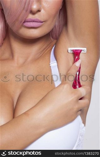 Attractive pink hair young woman gently shaving armpit epilation hair removal with ergonomic pink shaver. Body care skincare beauty concept