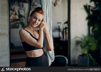 Attractive red haired woman in black sportswear stands by hammock and relaxing during antigravity yoga training in cozy ambiance of fitness center. Healthy lifestyle and wellness concept. Pleasant looking red haired fitness woman in black sportswear resting after aerial yoga class