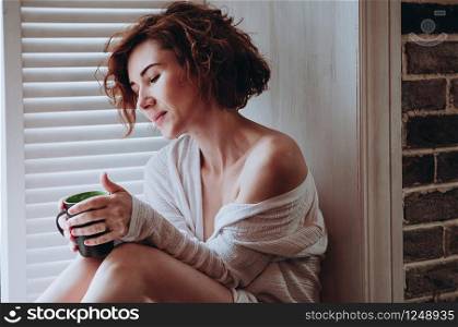 Attractive red-haired woman in white blouse, bear shoulders and legs, sitting on the window, holding a cup. Morning at home, relaxation time. Erotic, sensual, lifestyle concept. Soft focus image