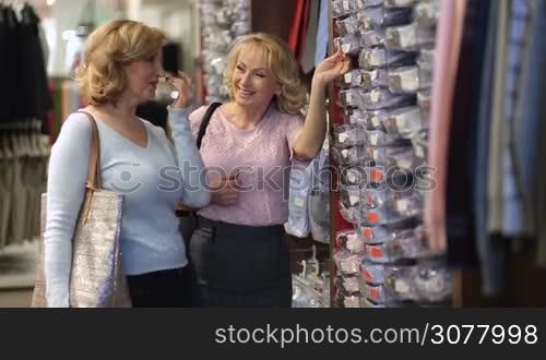 Attractive senior female friends with shopping bags looking for the right size of male shirt in the shelf during garments shopping in clothing store. Beautiful mature women choosing man shirt in fashion boutique shop.