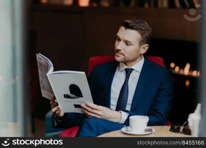 Attractive serious businessman dressed in formal suit, reads popular magazine, sits in coffee shop, drinks aromatic cappuccino, waits for business partner, focused in menu. People and rest concept