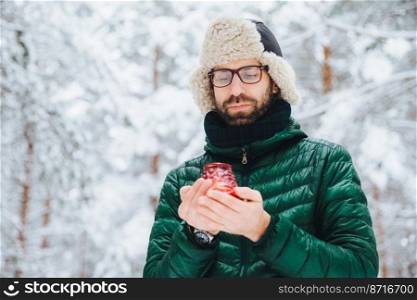 Attractive serious male wears warm winter clothes, keeps candle, stands against winter forest background, enjoys fresh air, being very confident. Fashionable unshaven man outdoors.