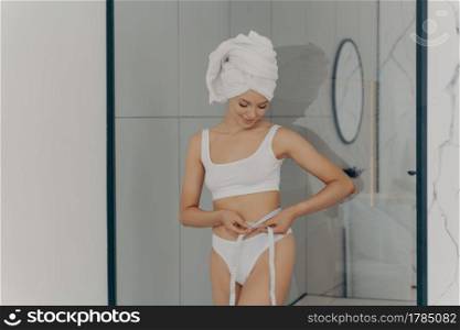 Attractive slender fit woman measuring her waistline with centimeter tape while standing in bathroom after shower in white classic underwear with hair wrapped in towel. Weight loss and dieting concept. Attractive slender fit woman measuring her waistline with centimeter tape