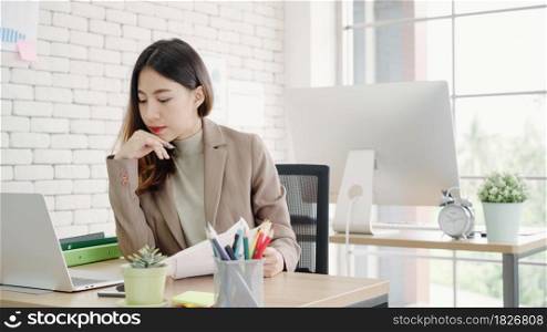 Attractive smart Asian business woman in smart casual wear working on laptop while sitting on desk while her angry manager standing angry throwing papers on office desk. Women work at office concept.