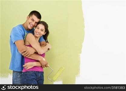 Attractive smiling couple posing in front of partially painted wall holding paint roller.