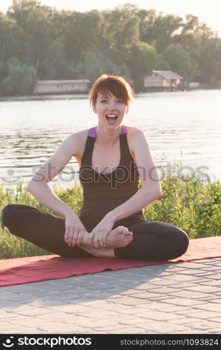 Attractive smiling female sitting in lotus yoga position, wearing tight black clothes, hands on bare feet, stretching pose, outdoor, water in background. Healthy lifestyle, keep fit, weight loss concept