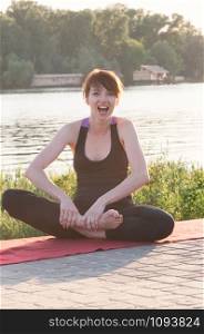 Attractive smiling female sitting in lotus yoga position, wearing tight black clothes, hands on bare feet, stretching pose, outdoor, water in background. Healthy lifestyle, keep fit, weight loss concept