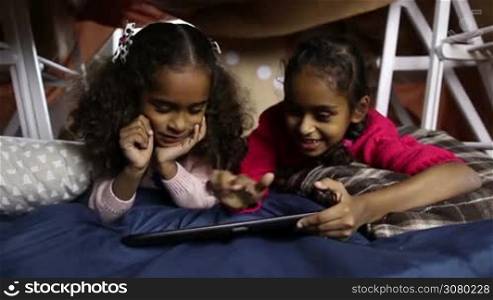 Attractive smiling little girls working on digital tablet pc while relaxing together at home in cubby house made of blanket and chairs. Positive mixed race sisters surfing the net on touchpad and smiling. Dolly shot. Slow motion.