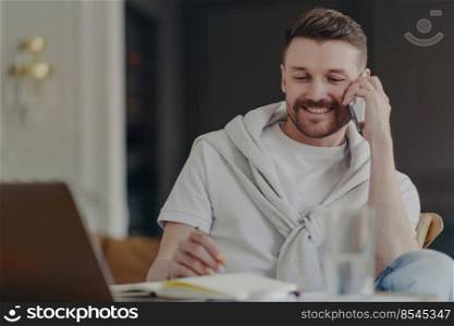 Attractive smiling male freelancer or businessman talking on phone while sitting at his work desk in front of laptop at home office making notes, wearing tshirt and sweater over his shoulders. Smiling freelance worker talking on phone while sitting at his work desk at home office