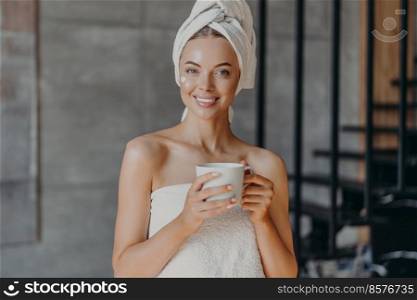 Attractive smiling young woman with makeup, toothy smile, poses bare shoulders, wrapped in towel, drinks hot beverage stands indoor has cheerful expression applies moisturizing cream on face