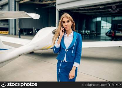 Attractive stewardess in uniform poses against small airplane in hangar. Air hostess in suit near plane. Private airline, flight attendant. Stewardess in uniform poses against small airplane