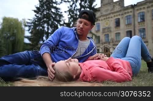 Attractive student couple relaxing on campus lawn after lesson. Man sitting on the grass and touching gently woman beautiful blond hair with tenderness and chatting while cute girl lying on the back and looking at blue sky.
