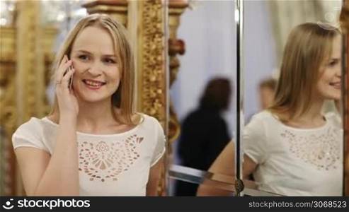 Attractive stylish woman using a mobile phone reflected in a large mirror alongside her in luxury indoor surroundings