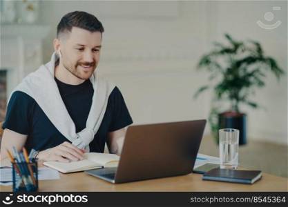 Attractive successful male freelancer wearing earphones taking part in online meeting while sitting in front of laptop at his cozy workplace at home or office, manager in casual wear sitting behind office desk. Happy businessman having online meeting, looking at laptop while working at home
