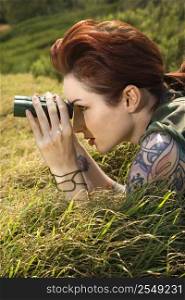 Attractive tattooed Caucasian woman lying on grass looking through binoculars in Maui, Hawaii, USA.